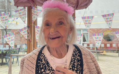 Smiling 100 year old Pamela Bankes wearing a pink party hat and with birthday bunting decorating the large windows in the background. 