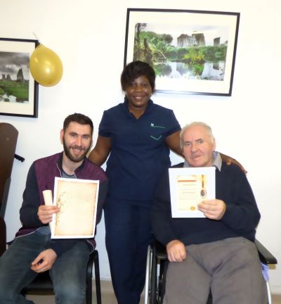 Two seated male residents with their 'celebrating resilience certificates' at Cashel Residential Older People Services. Standing behind them is a nurse with a supporting hand on both of their backs. 