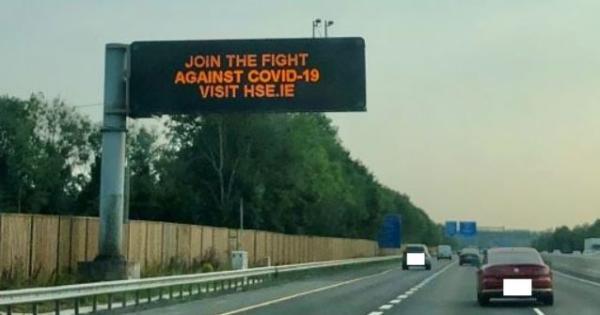 Photograph of a big sign on the side of the M7 motorway saying JOIN THE FIGHT AGAINST COVID-19 - VISIT HSE.ie with cars driving past