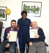 Two seated male residents with their 'celebrating resilience certificates' at Cashel Residential Older People Services. Standing behind them is a nurse with a supporting hand on both of their backs