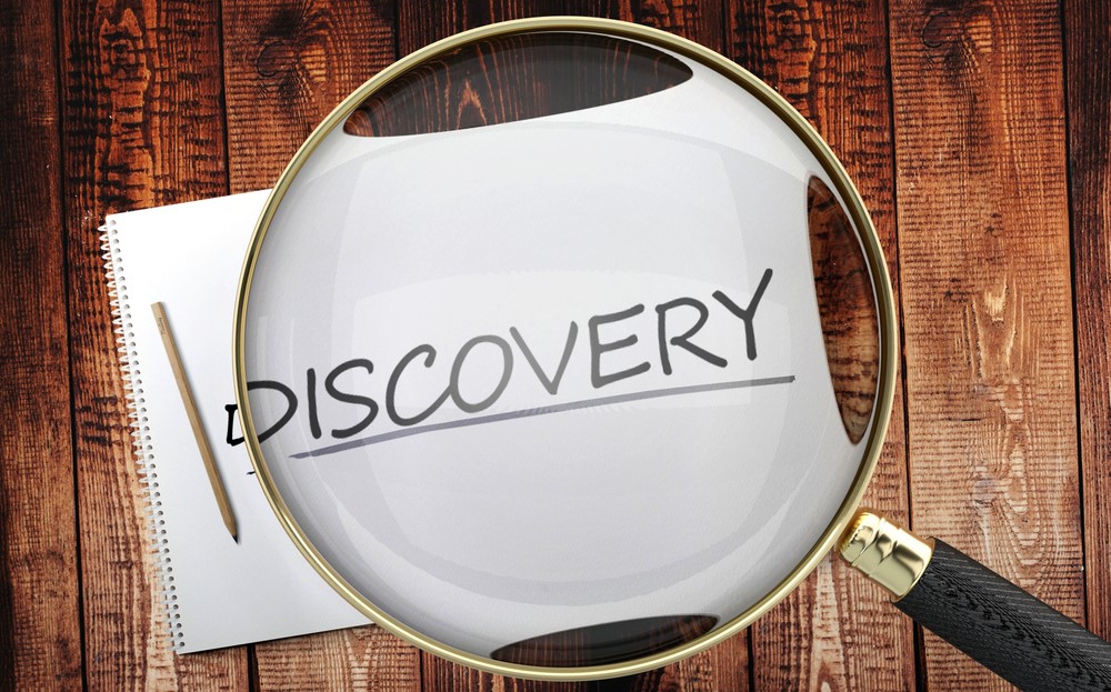 A magnifying glass is held over the word 'discovery' which is written in capital letters on a blank white sheet, lying on a board.