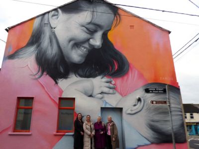 Four women standing outdoors in front of a large mural of a woman breastfeeding a baby.