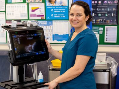 A woman in medical clothes stands beside an ultrasound machine in a clinical setting.