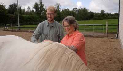 A woman in a pink shirt and a young man in a grey shirt are standing beside a horse. They are patting the horse.