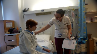 A female patient is seated on a bed in a hospital. A health care assistant is leaning forward and touching the patient's shoulder.