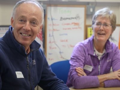 A man and woman sitting at a desk indoors and smiling.