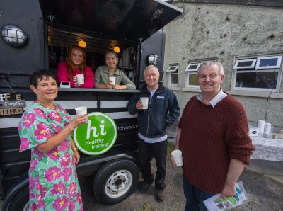 3 people, 2 men and a woman standing before a coffee tricks with coffee cups in their hands. A healthy Ireland logo is on the coffee truck and 2 women are inside the coffee truck. 