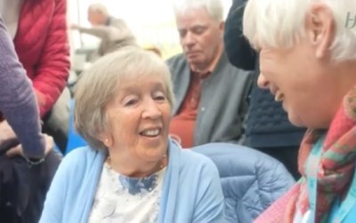 A lady is sitting down. She is wearing a blue cardigan. She is smiling up at a lady who is standing and smiling back at her.