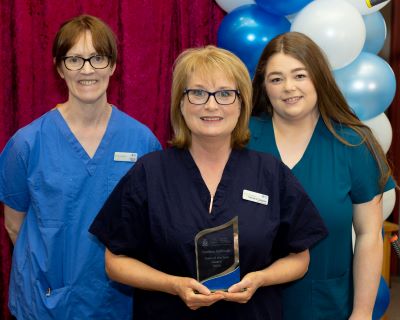 3 female healthcare professionals standing in a row. The lady in front is holding an award. There are blue and white balloons in the background. 