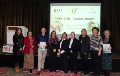 Large group of people standing in a row in front of a large screen that reads 'Hey You - Listen, Hear.