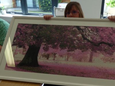 A woman sitting with a framed painting of some trees on her lap.