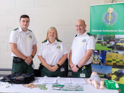 Three National Ambulance Service staff standing behind a table with first aid material on it, beside them is a National Ambulance Service pull-up poster.