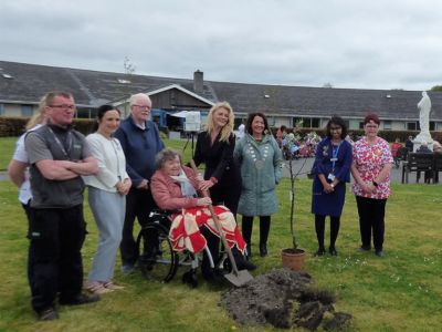 Eight people stand outside on a grassy lawn in front of a large building. An older woman in a wheelchair beside a potted baby tree holds a shovel.