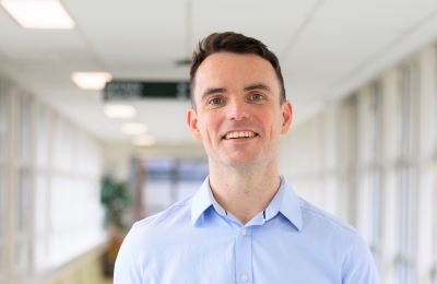 Dr Adam Dyer smiling. He is wearing a blue shirt and is standing in a hospital corridor. 