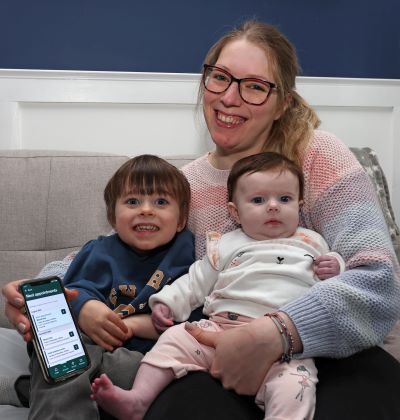 A smiling woman is seated holding two young children; a little boy and a baby in her arms. She is holding a phone with teh HSE Health App displayed on it. 