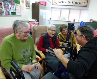 3 ladies are seated and laughing while a man in a black hoody is knelling before them. 