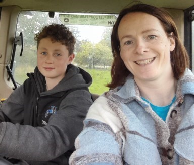 A boy is seated in a tractor beside a smiling woman. The boy has his hands on the steering wheel. 