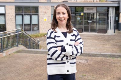 Gemma Donegan standing outside a healthcare building. Gemma is standing with her arms folded. She is wearing a black and white striped cardigan. 