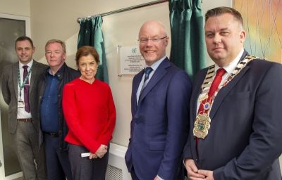 Five people standing in front of a plaque on a wall, there are green curtains either side of the plaque. 