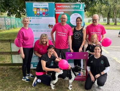 Group of 9 people, including a child wearing running pink and black running gear. They are standing beside a pull up stand that reads 'maternal sepsis. 