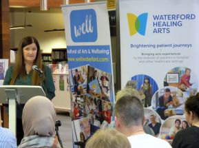 Woman standing a lectern in front if a group of people. There are 2 pull up banners beside the woman, one reads Festival of Arts and Wellbeing. The second one reads, Waterford Healing Arts