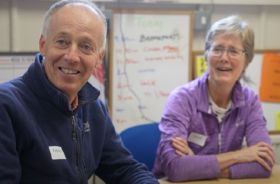 A man and woman sitting at a desk indoors and smiling.