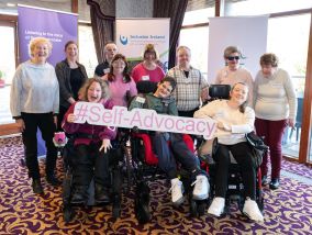 Large group of staff and service users in a function room for the “Hear My Voice” Self-Advocacy Event. 3 wheelchair users  are seated in the front row. Together they are holding a sign that reads: #Self-Advocacy 