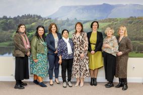 Group of 8 women standing in a row in front of a scenic backdrop