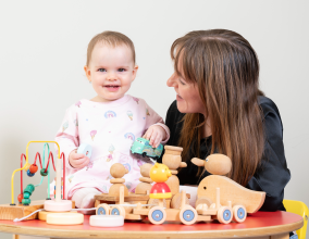 A smiling baby girl is sitting on a table surrounded by wooden toys. She is supported by a women with long hair who is looking at the child fondly.