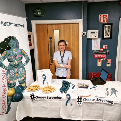 Natasha Mahon standing behind a table at an information stand. The signage reads #choosescreening 'Eliminate Cervical Cancer'. 