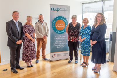 A group of 6 people standing in aroom on eaither side of a National Cancer Control Programme pull up stand. 